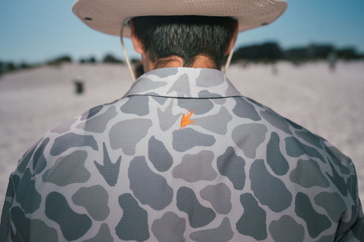 Person wearing a camouflage shirt with a frog foot logo, standing on a beach.