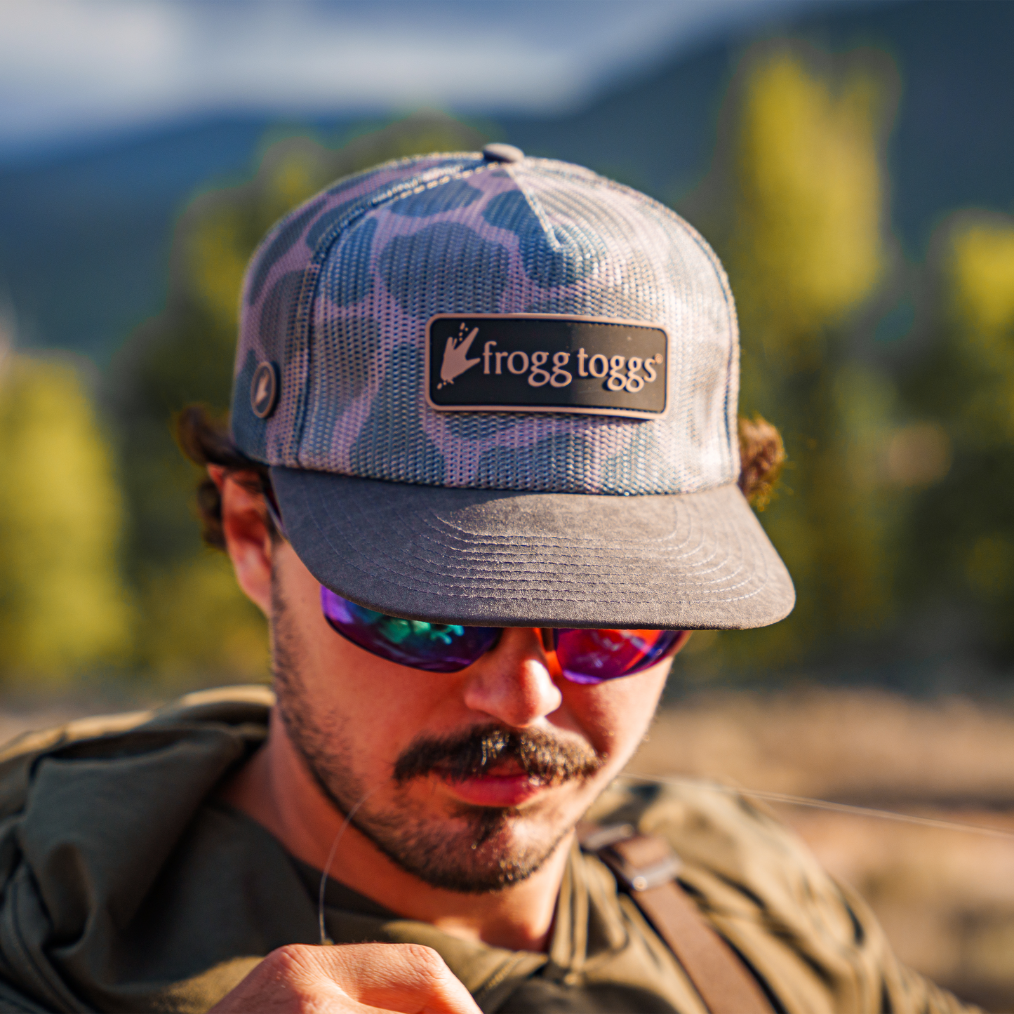 A man outdoors wearing a gray and blue patterned trucker hat with a brown patch that reads 'frogg toggs' and reflective sunglasses, with a blurred natural background.