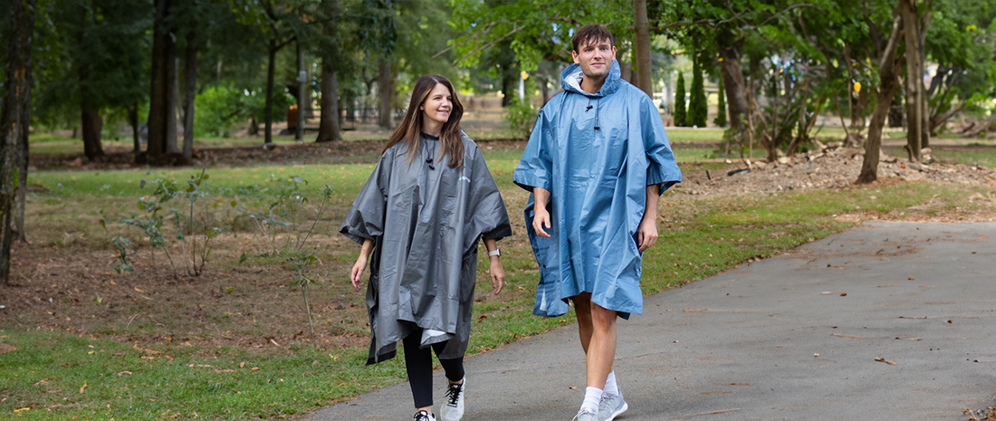 two people walking in a park on an overcast day wearing frogg toggs ponchos. Man in blue poncho, woman in black poncho.