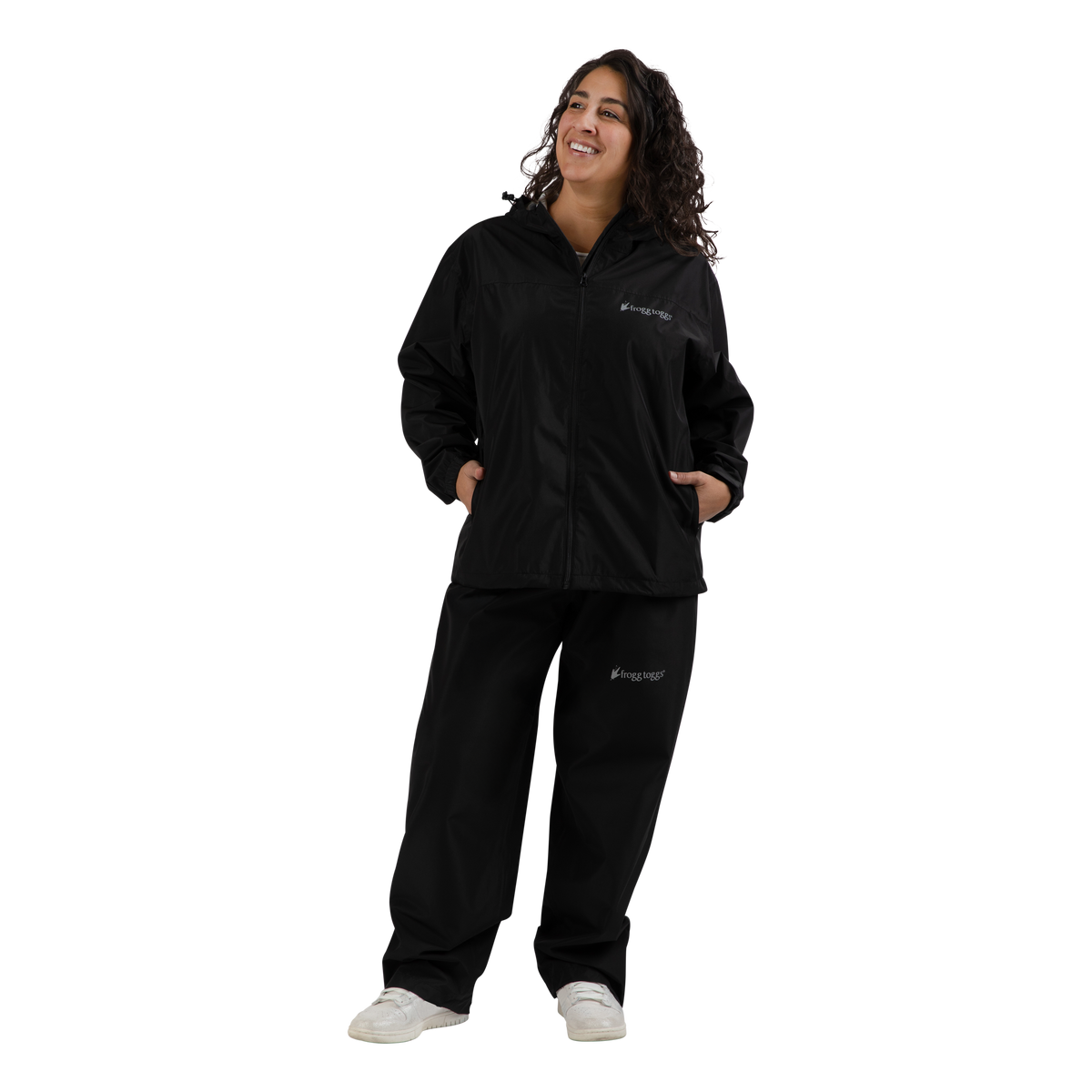A woman modeling a black zip-up jacket and matching black sweatpants set with small white 'frogg toggs' logos on the chest and left thigh, paired with white sneakers, against a solid white background.