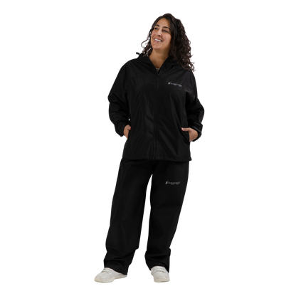 A woman modeling a black zip-up jacket and matching black sweatpants set with small white 'frogg toggs' logos on the chest and left thigh, paired with white sneakers, against a solid white background.