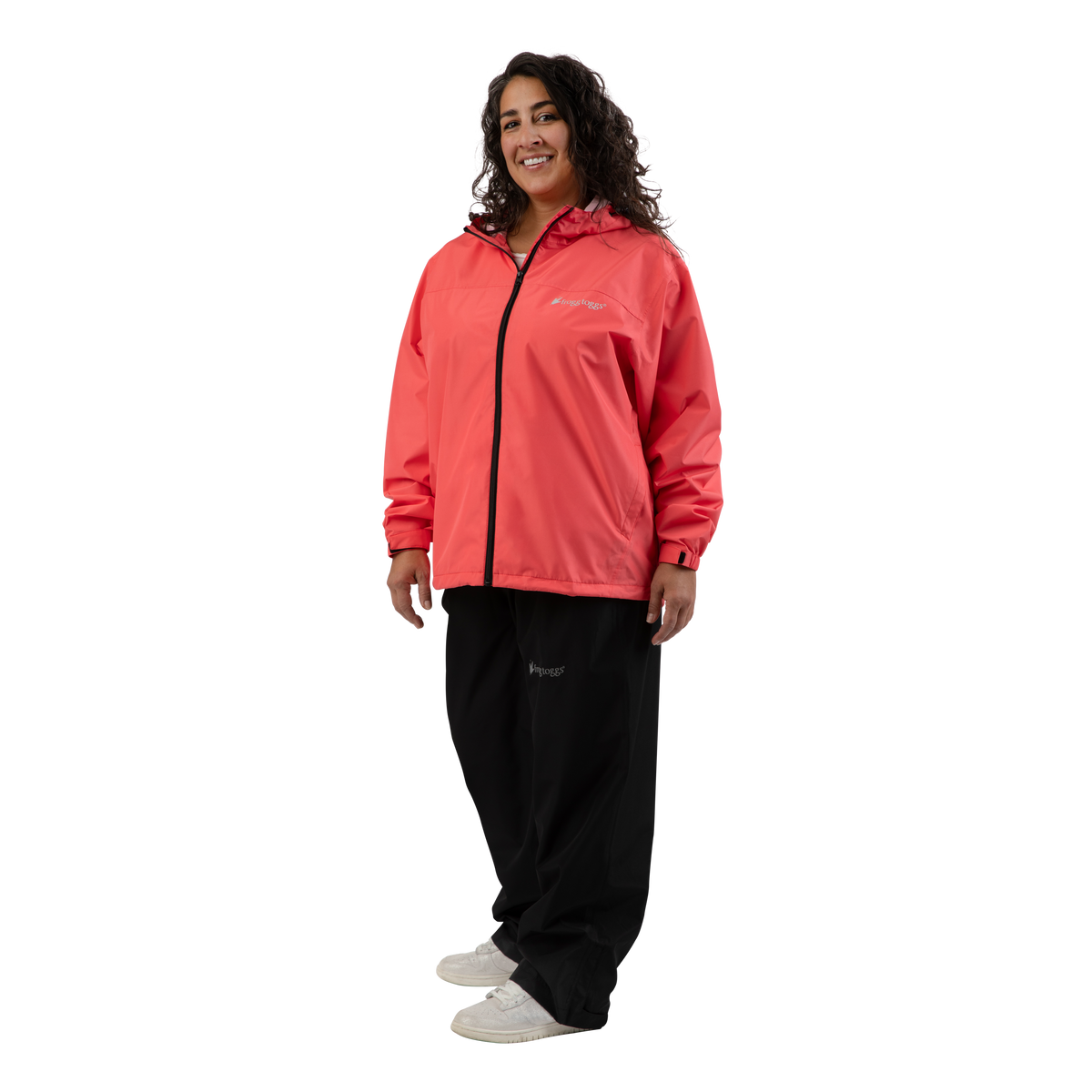 A woman modeling a coral pink hooded zip-up jacket with black zippers and a small white frogg toggs logo on the chest, paired with black sweatpants and white sneakers, against a solid white background.