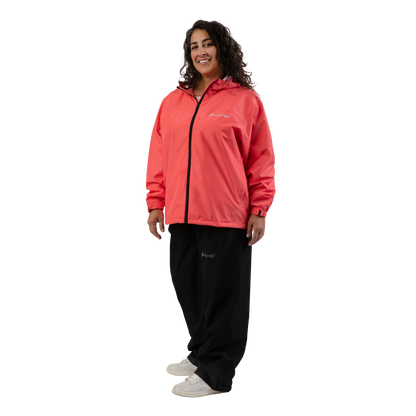 A woman modeling a coral pink hooded zip-up jacket with black zippers and a small white frogg toggs logo on the chest, paired with black sweatpants and white sneakers, against a solid white background.