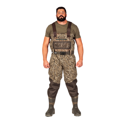A man modeling a pair of camouflage chest waders with brown suspenders, an olive green t-shirt, and dark rubber boots, set against a plain white background.