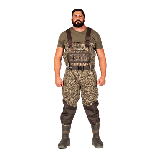 A man modeling a pair of camouflage chest waders with brown suspenders, an olive green t-shirt, and dark rubber boots, set against a plain white background.