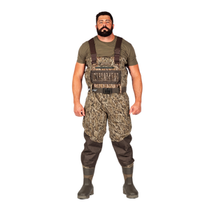 A man modeling a pair of camouflage chest waders with brown suspenders, an olive green t-shirt, and dark rubber boots, set against a plain white background.