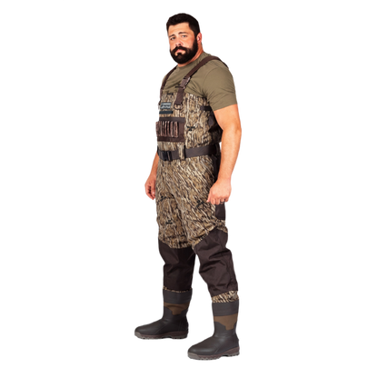 A man wearing a pair of camouflage chest waders over an olive green t-shirt and black rubber boots, standing against a solid white background.