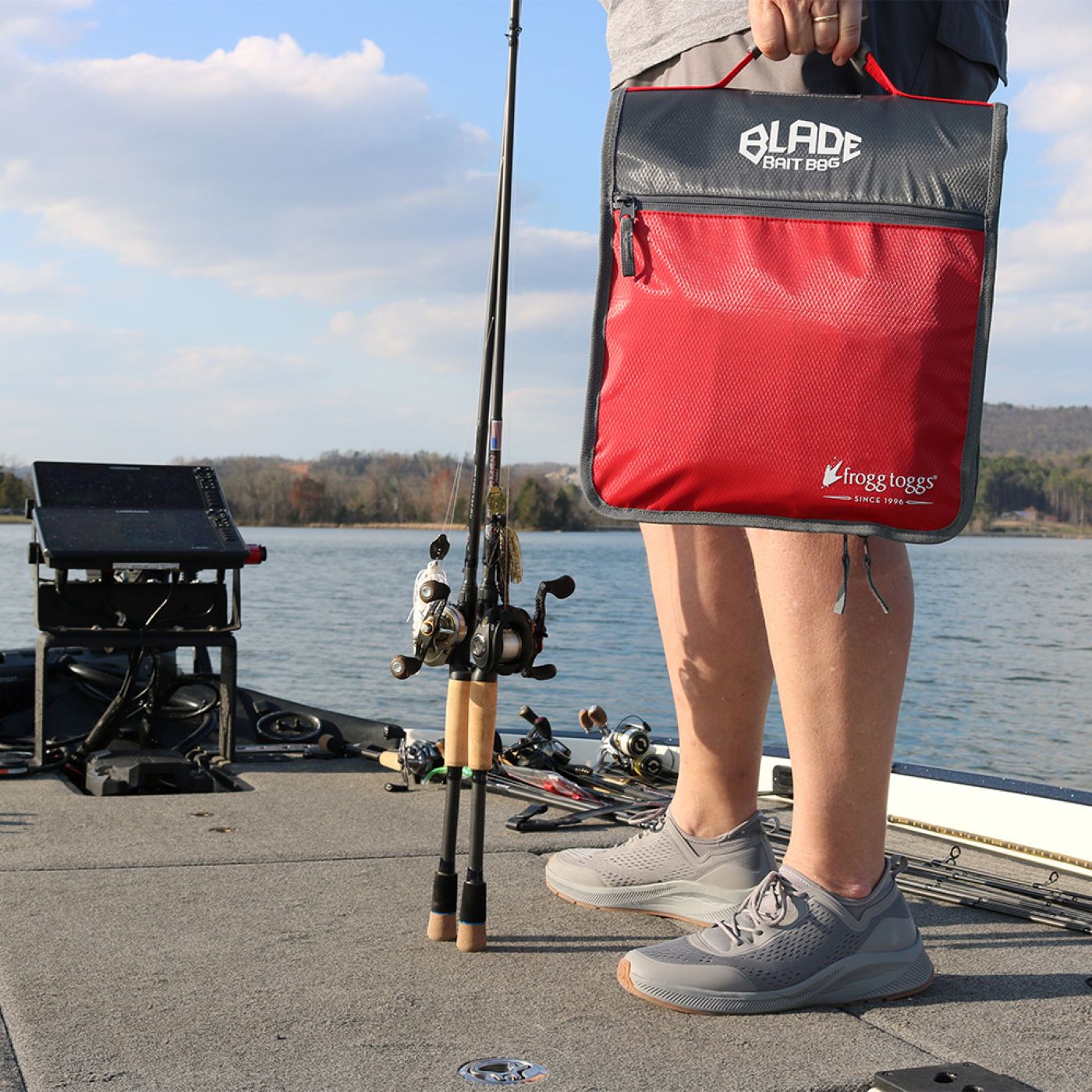 Person holding a red and black fishing tackle bag by a lake with fishing equipment.