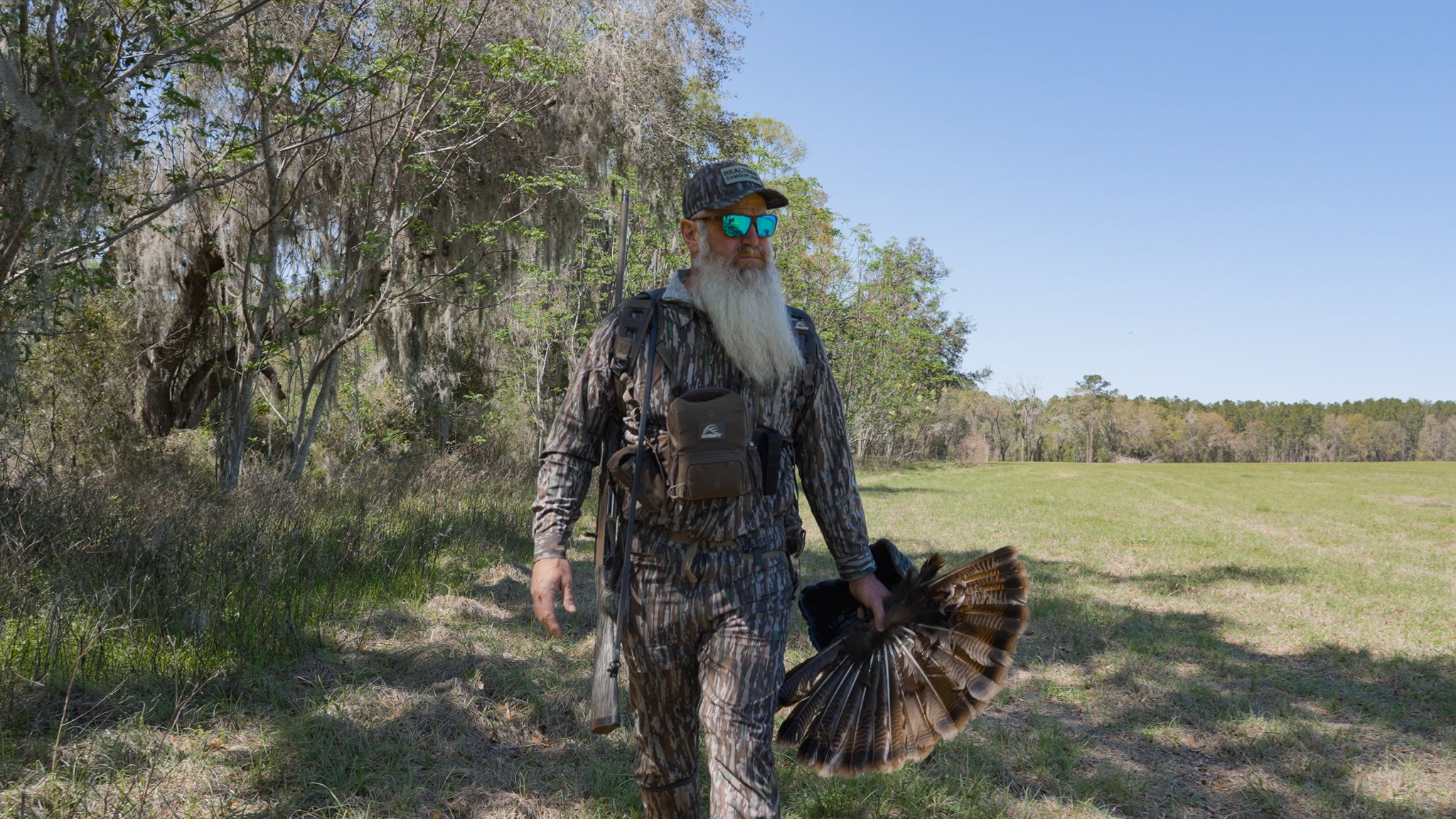 Man in camouflage walking outdoors with a turkey in his hand