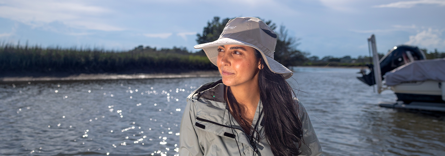 Woman wearing a hat and jacket standing by a body of water with a boat in the background.