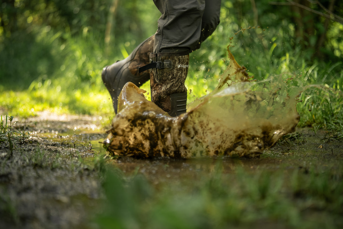 A person wearing muddy Frogg Toggs boots and dark pants walks through a muddy puddle on a forest path, creating a large splash of wet mud and grass.