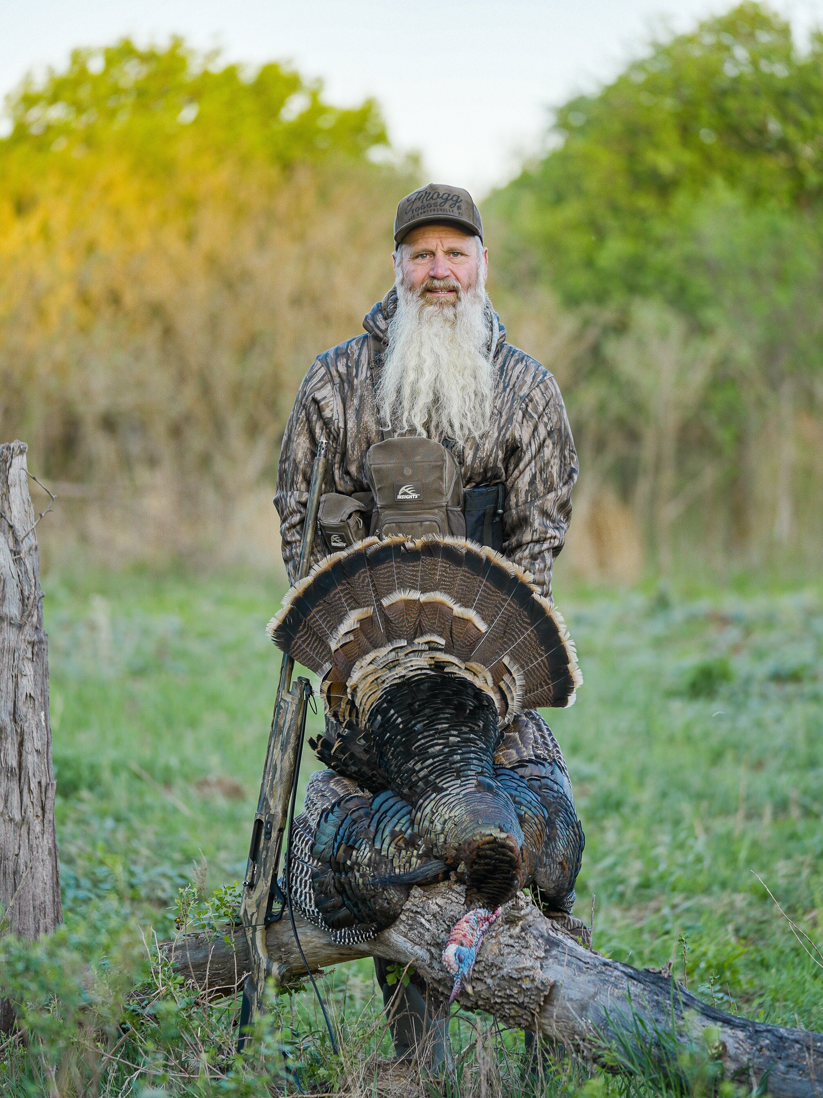 Man with a long beard holding a large turkey in a field with trees in the background