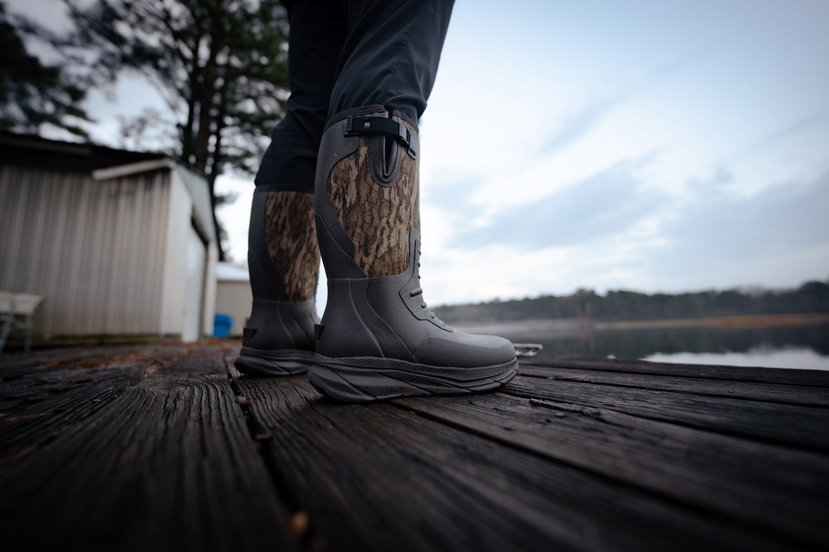 Gray waterproof Frogg Toggs hunting boots with brown camouflage knit leg panels, worn by a person standing on a wooden dock outdoors near water and trees.