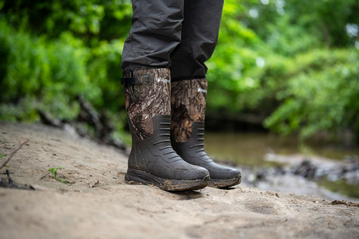 A person standing outdoors near a stream wearing dark gray Frogg Toggs waterproof pants and camouflage-patterned rubber boots with dark gray soles.