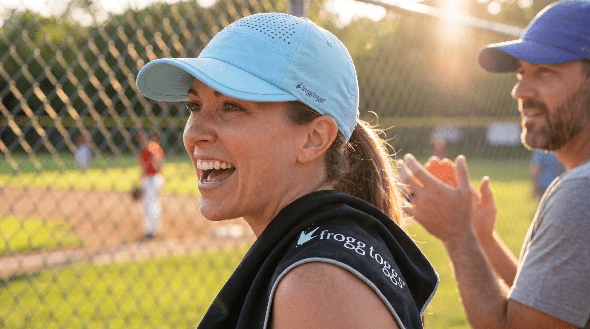 Woman in a blue cap and black sleeveless shirt with 'frogg toad' logo on a sports field, clapping hands with a man.