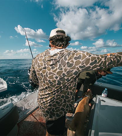 Man in camouflage shirt and cap on a boat with a dog, looking out at the ocean under a blue sky with clouds.