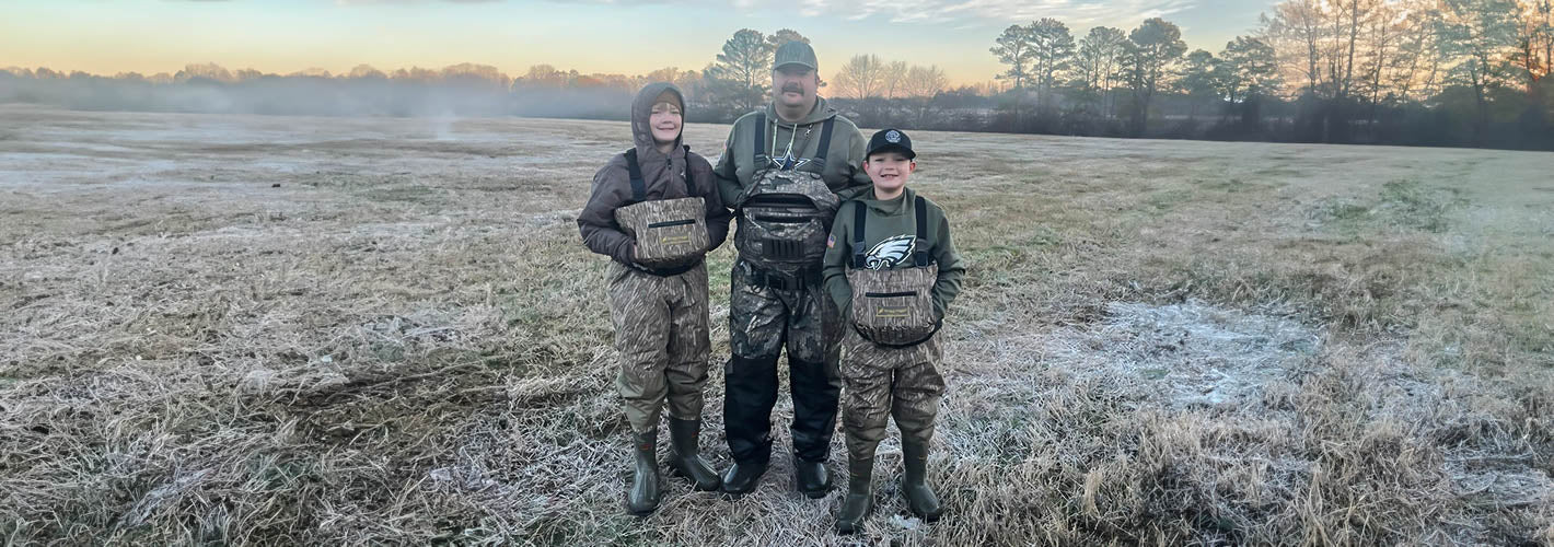Three people in camouflage clothing standing in a field with a sunset or sunrise in the background.