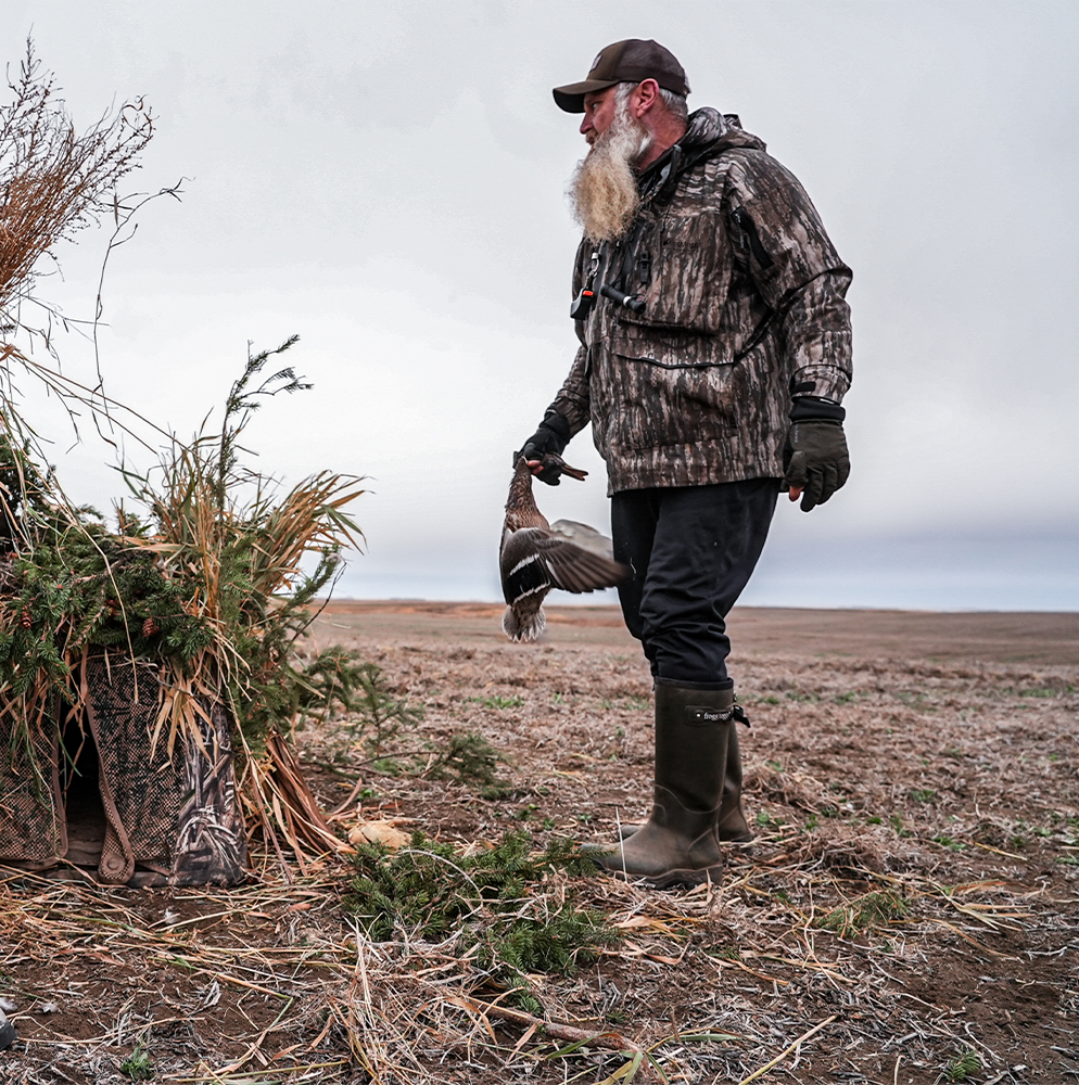 Man in camouflage hunting gear holding a duck in a field