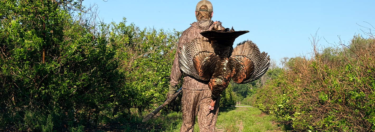 Hunter in camouflage clothing with a large bird, likely a turkey, in a natural setting.