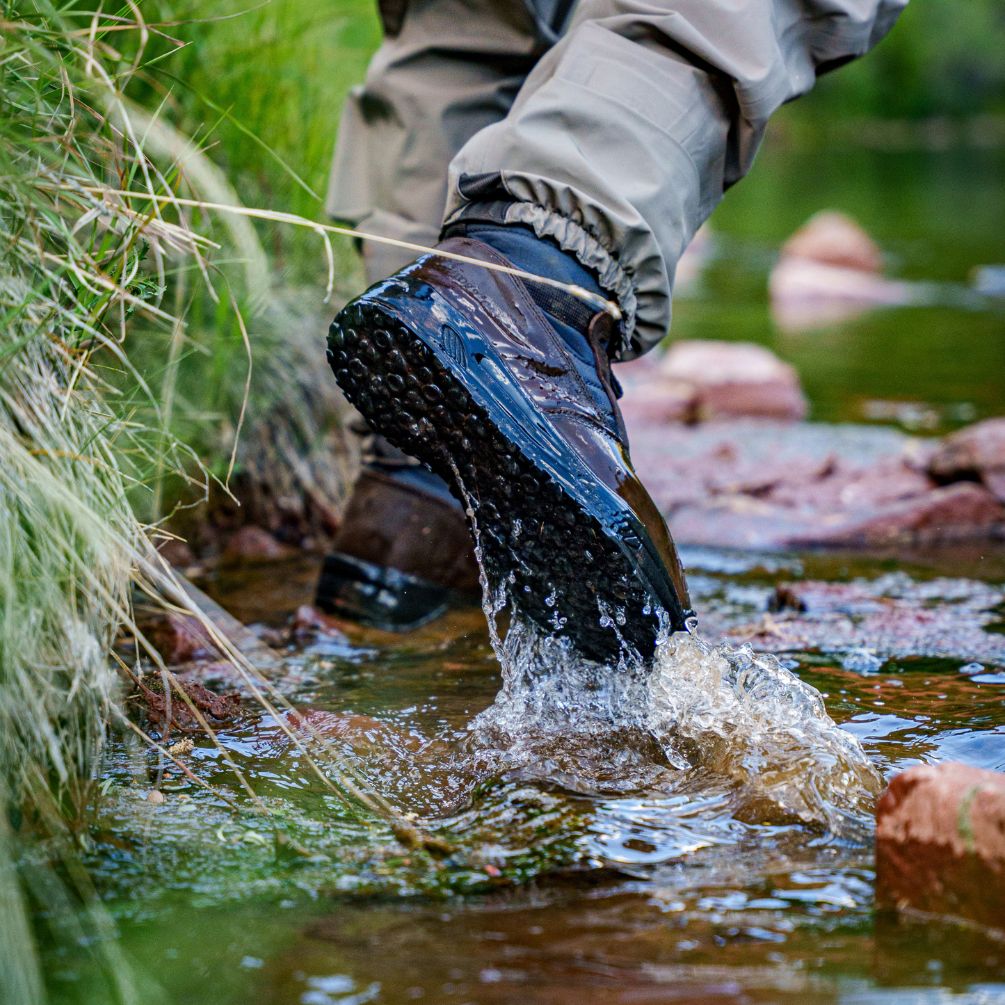 A close-up outdoor shot of a person's legs and Frogg Toggs boots stepping into a shallow stream, with water splashing around the sole of a dark waterproof hiking boot. The person is wearing khaki pants and the setting includes grass, rocks, and green water.