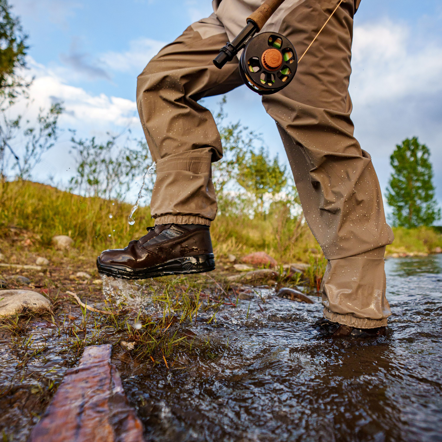 A person wading through shallow water in a river, wearing tan waterproof Frogg Toggs fly-fishing pants and dark brown Frogg Toggs boots, with a fly fishing rod hanging from the waist.