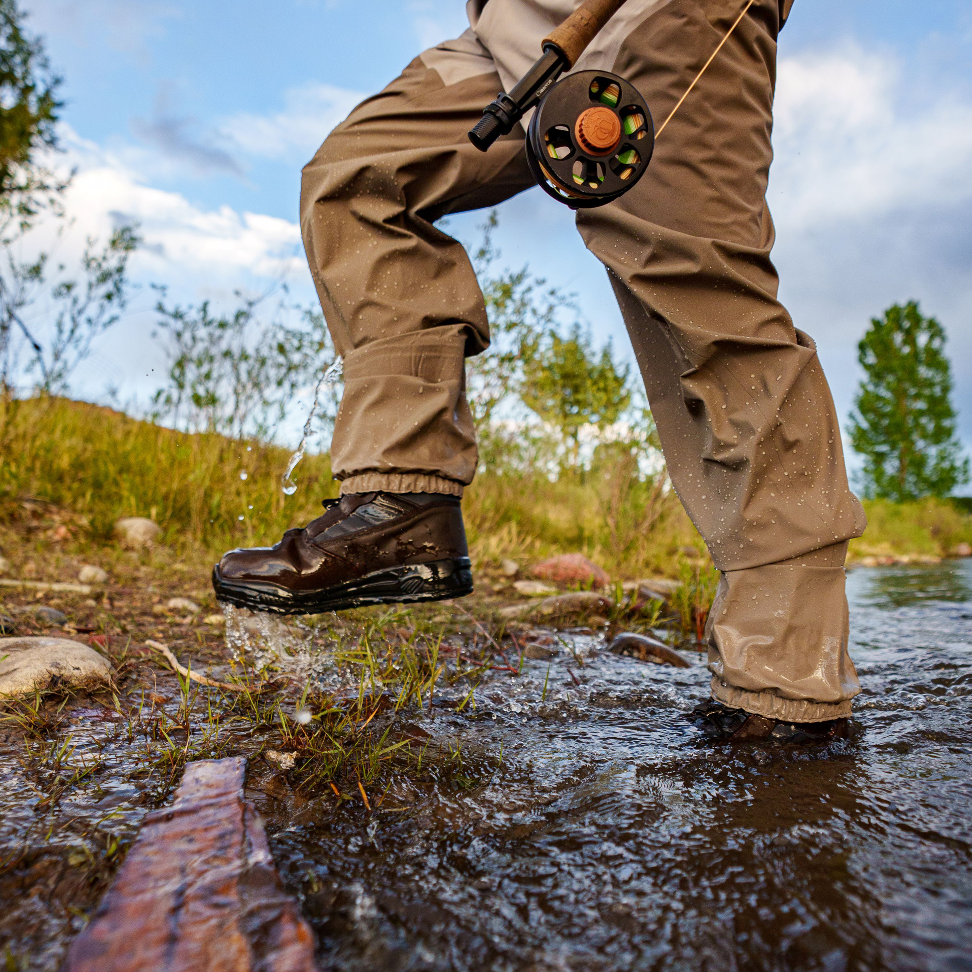 A person wading through shallow water in a river, wearing tan waterproof Frogg Toggs fly-fishing pants and dark brown Frogg Toggs boots, with a fly fishing rod hanging from the waist.