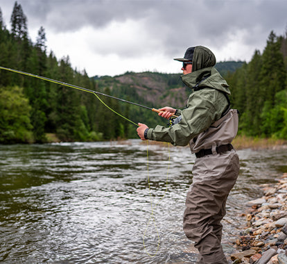 A person wearing a green hooded rain jacket and sunglasses stands by a river in the rain, holding a fly fishing rod and a black chest-mounted bag with fishing gear inside. Trees and rocks line the riverbank.