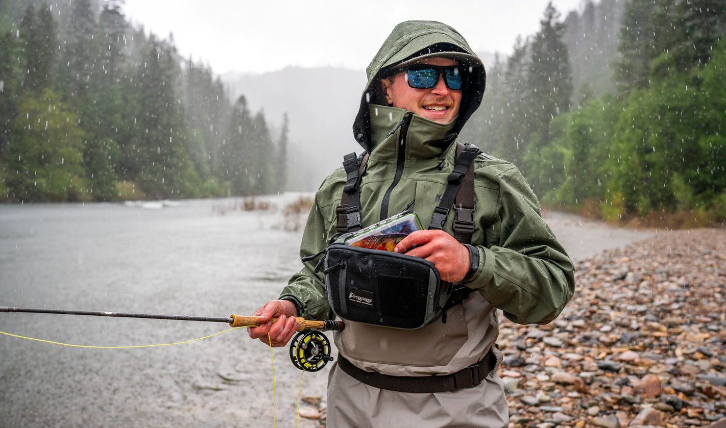 A person wearing a green hooded rain jacket and sunglasses stands by a river in the rain, holding a fly fishing rod and a black chest-mounted bag with fishing gear inside. Trees and rocks line the riverbank.