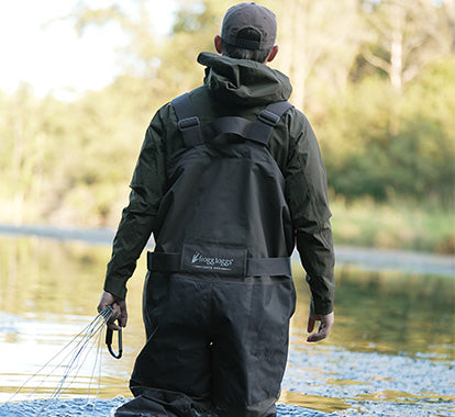 A close-up of a the back of a person wearing  frogg toggs waterproof waders over an olive green jacket, standing outdoors near water with a blurred natural background. Holding line and a hook
