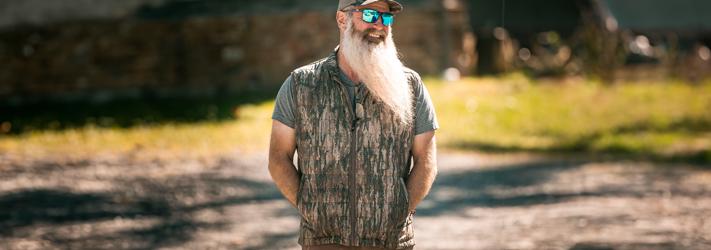 Man with a beard and sunglasses standing outdoors in a natural setting