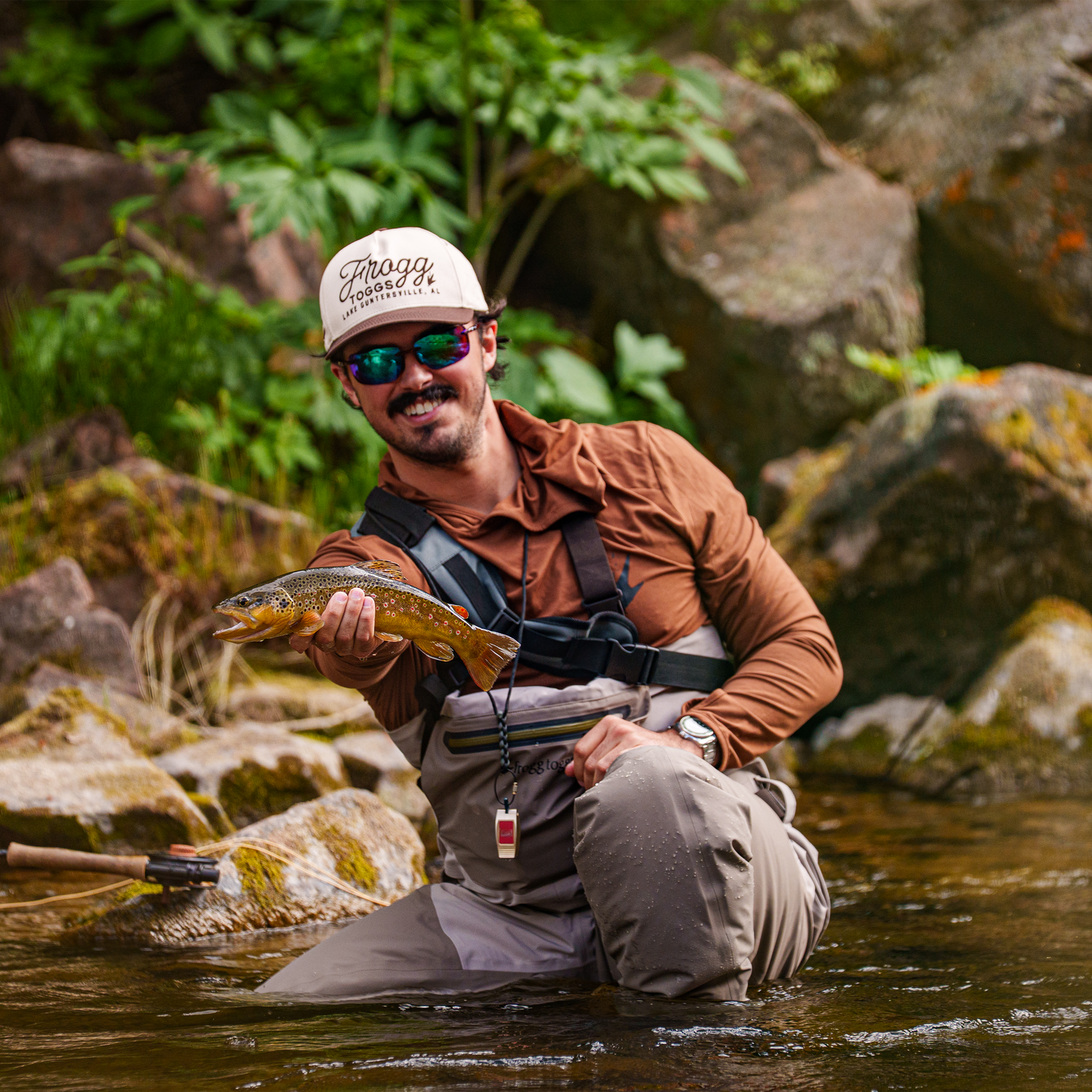 Man holding a fish in a river with rocks and greenery