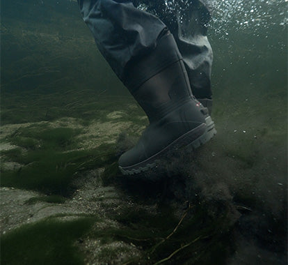 A person wearing dark gray waterproof boots and pants steps into shallow, murky water, creating a splash of sediment and bubbles. The boots are black with a small red logo on the side.
