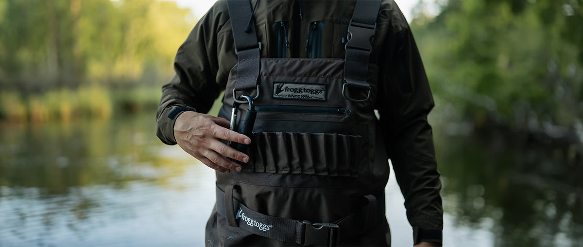 A close-up of a person wearing  frogg toggs waterproof waders over an olive green jacket, standing outdoors near water with a blurred natural background.